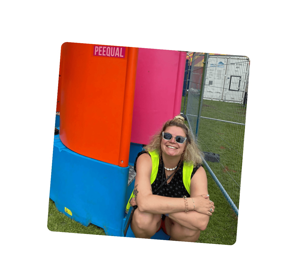 A happy blonde haired woman sitting in front of a colourful Peequal urinal setup, wearing a high-visibility vest and sunglasses at an outdoor festival location.